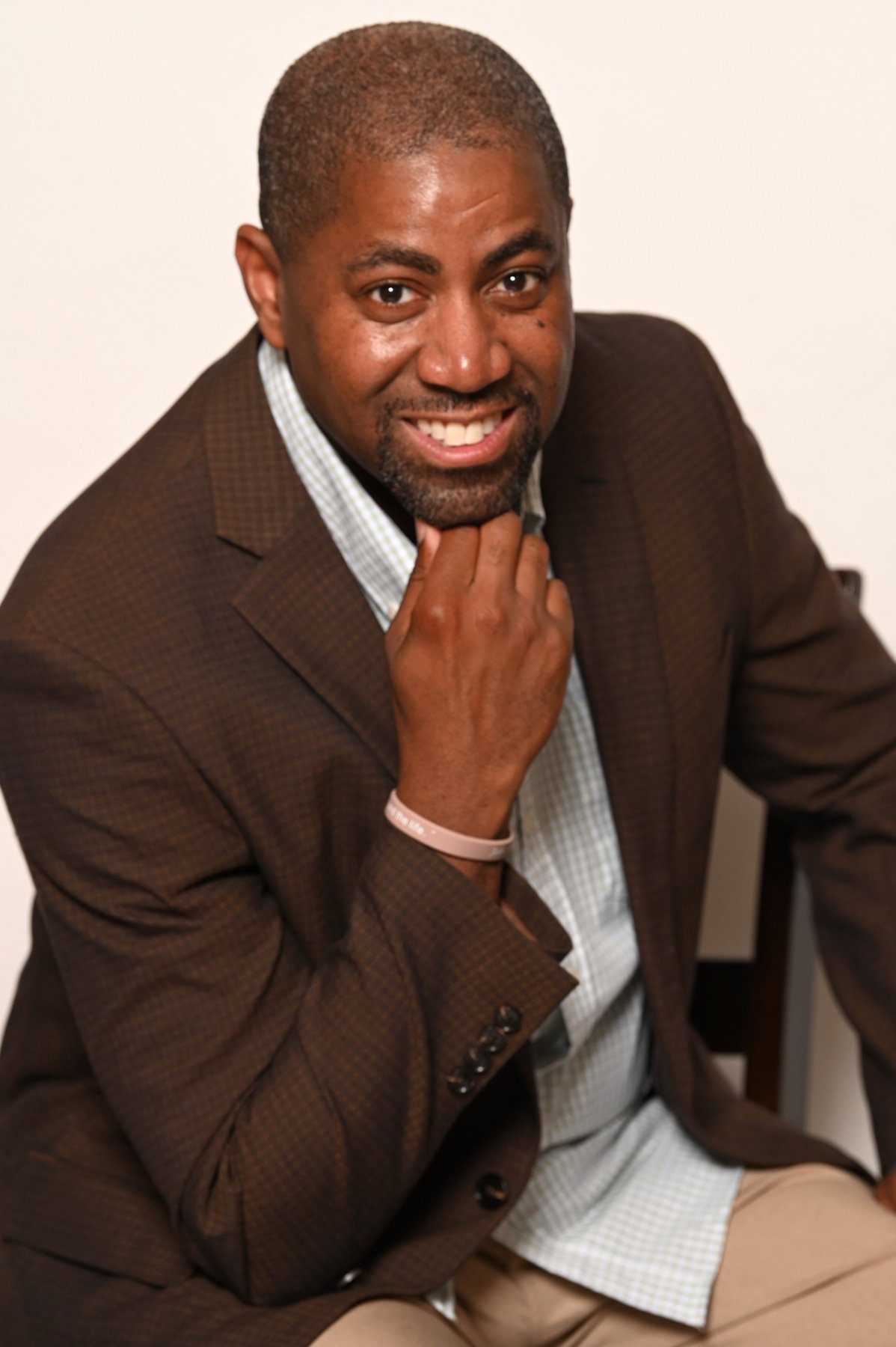 Pastor Larry D Smith, a middle-aged African American man wearing a brown checked suit jacket and light shirt, seated thoughtfully with his hand resting on his chin in a calm and welcoming indoor setting, conveying a sense of warmth and reflection